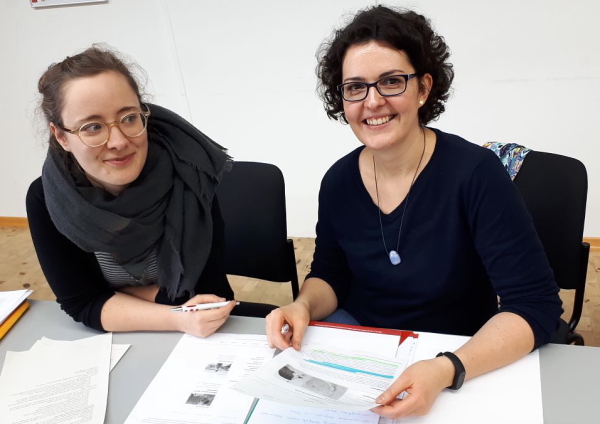 Aufnahme aus einem unserer Workshops: Zwei Frauen sitzen mit Unterlagen am Tisch. Eine schaut in die Kamera, die andere zur Seite. (Foto: ArbeiterKind.de) 